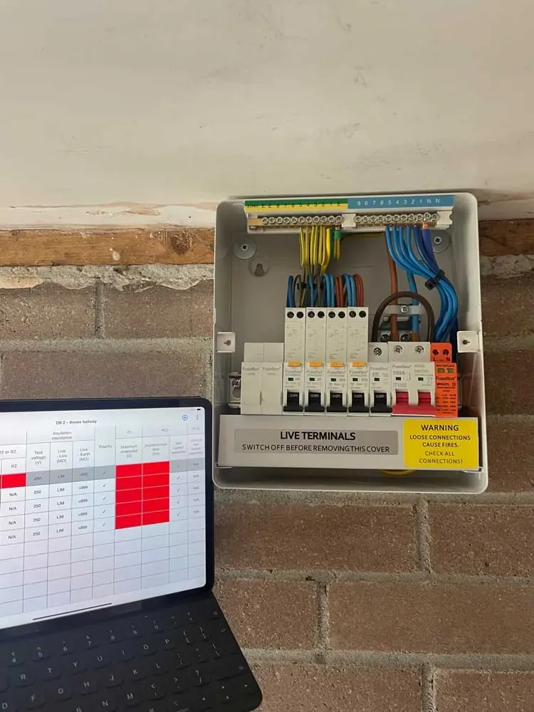 Wall-mounted consumer unit with its cover off during testing, insulation-resistance results being recorded on a tablet