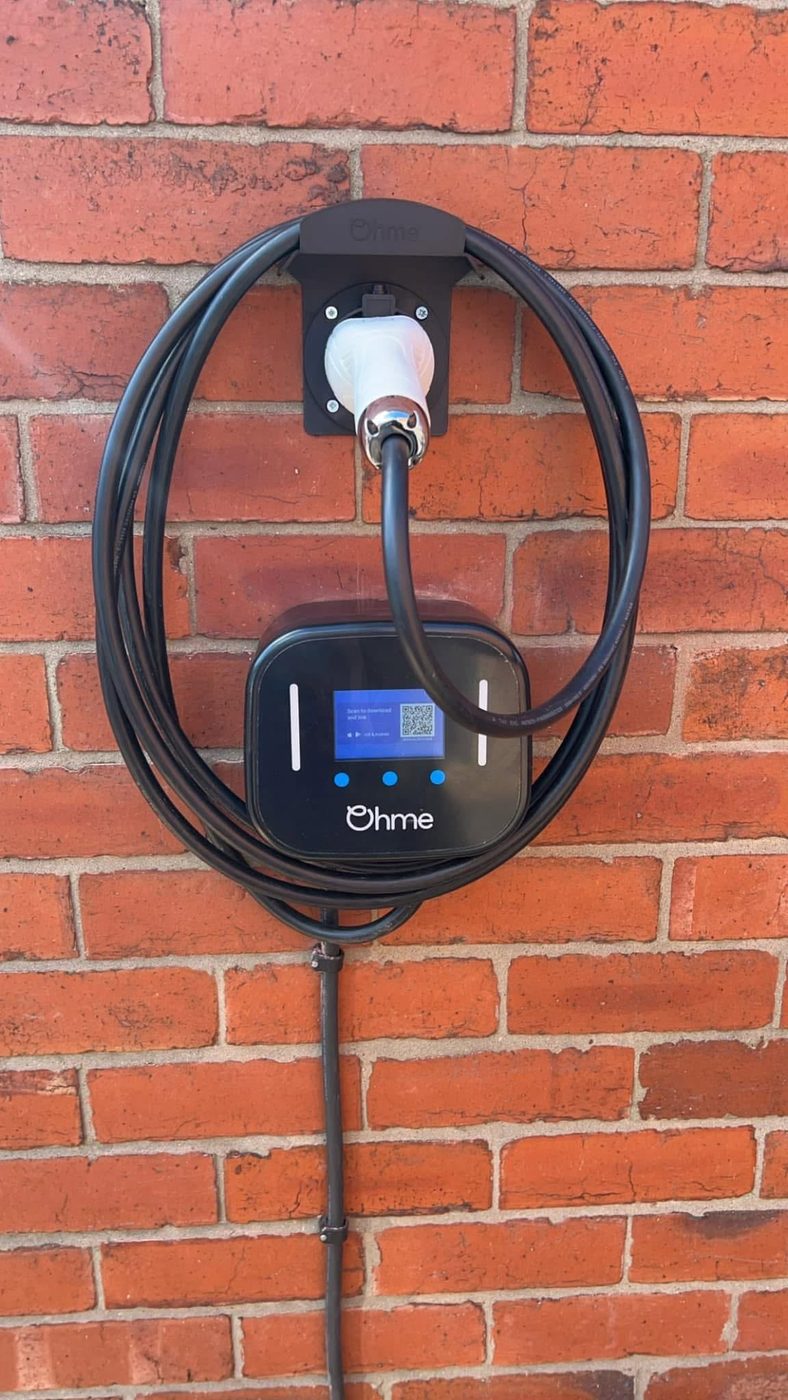 Ohme home EV charger installed on a red-brick external wall with the charging cable coiled on its holster